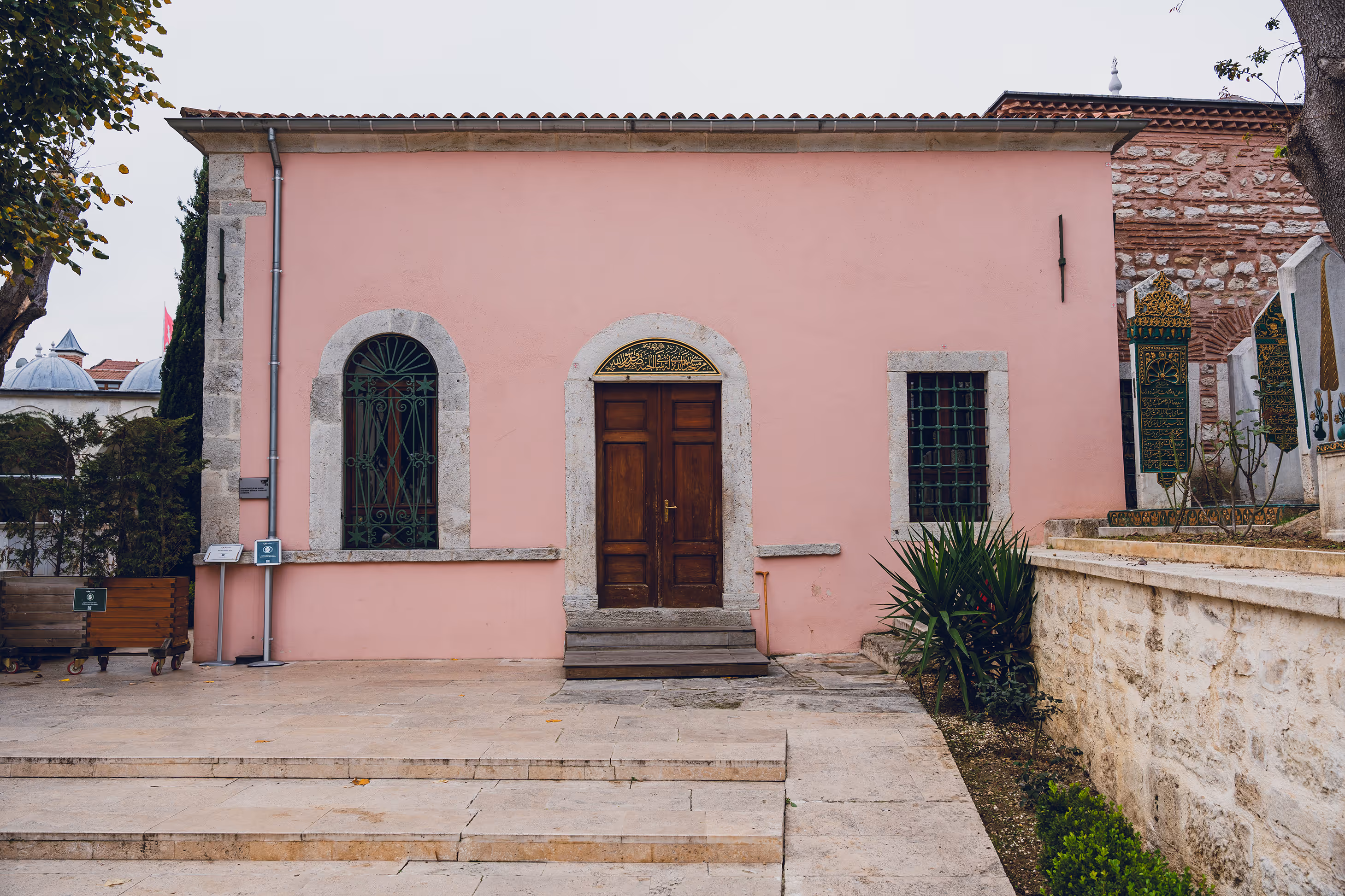 Pink-walled historic building with a wooden arched door, two barred windows, stone steps, and green plants nearby.