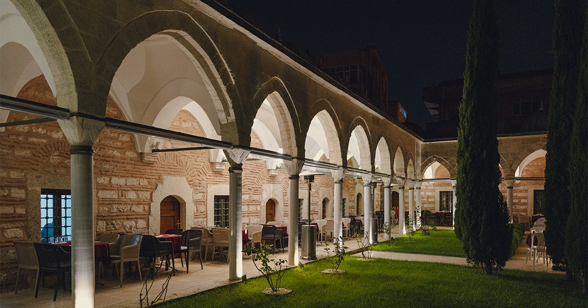Night view of an illuminated courtyard with arched colonnades, outdoor seating, and tall cypress trees.